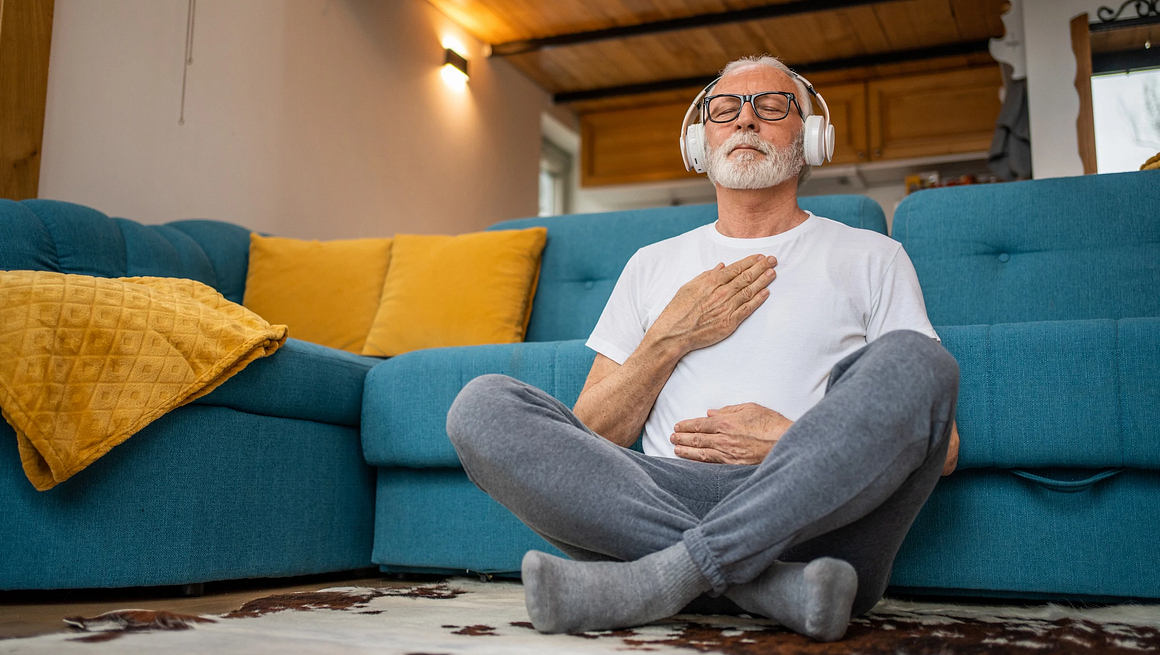 Mature senior man sitting on floor practicing guided meditation at home, relaxing body and mind concept