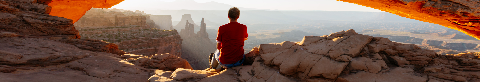 Person sitting beneath a rock arch overlooking a sunlit canyon landscape.