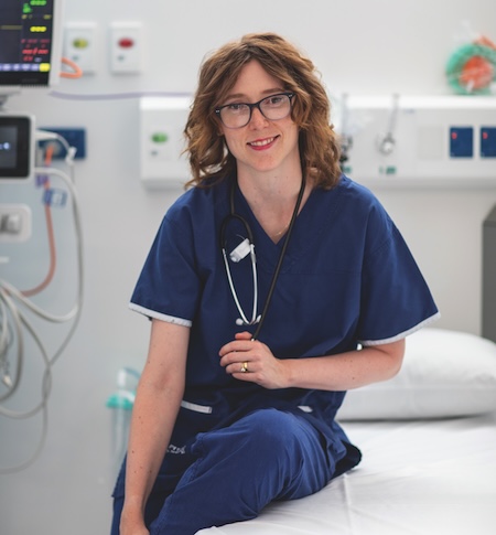 A woman wearing scrubs and stethoscope sitting on a hospital bed smiling into the camera